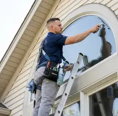 Professional window cleaner on a ladder using a squeegee to clean an exterior arched window, Mobile, AL.
